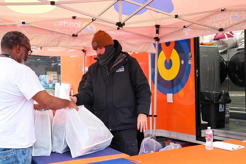 Two men stand under a tent next to an orange mobile laundry trailer as one hands white bags filled with clean clothes.