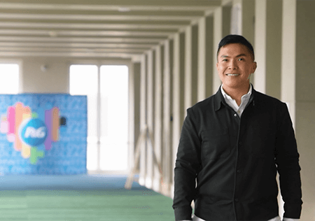 An Asian man stands in the hallways of his office and smiles at the camera.