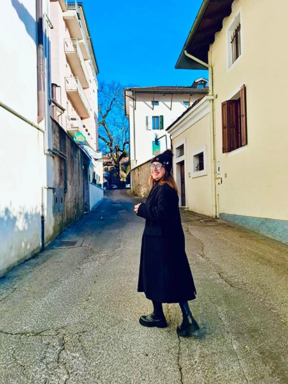 A woman wearing a long black coat, hat and sunglasses smiles while walking down a quiet, narrow paved street between buildings.