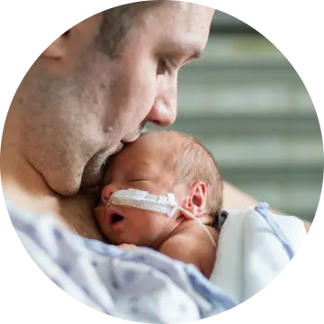 A man is holding a newborn baby on his chest. He is giving the baby a kiss on the forehead. The baby has a feeding tube taped to their cheek.