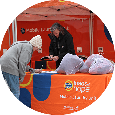 A woman volunteer stands at an orange Tide Loads of Hope table assisting another woman with laundry bags in cold weather.