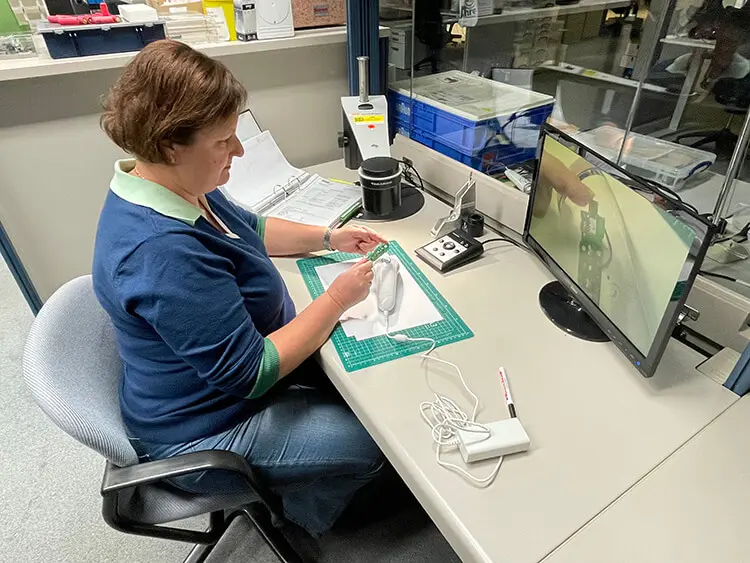 A woman with short, brown hair sits at her desk in a laboratory. She holds a small camera as she films the features of a handheld hair removal device.