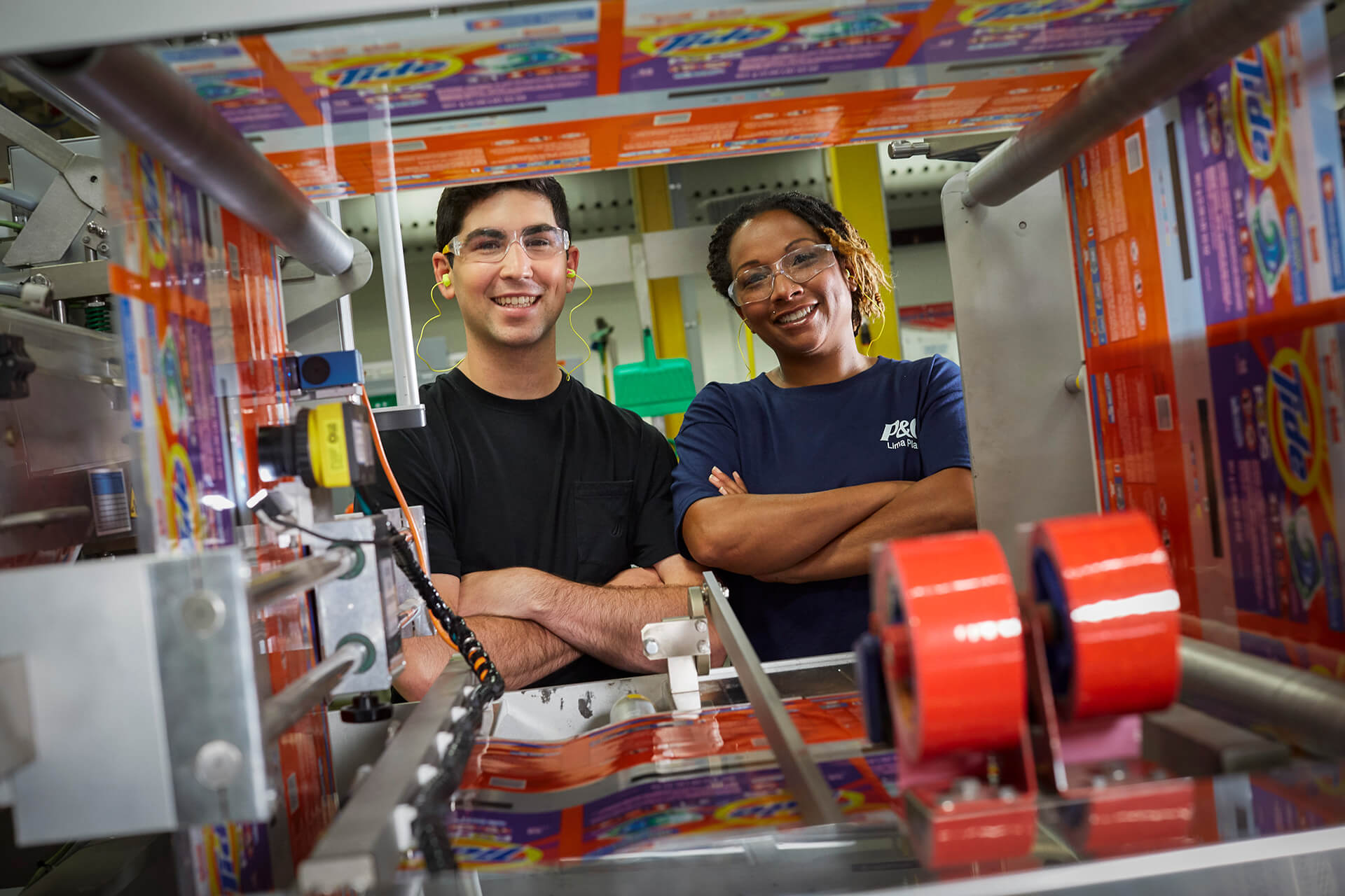 A male and female employee are in a manufacturing plant. They smile directly at the camera through some of the machinery.