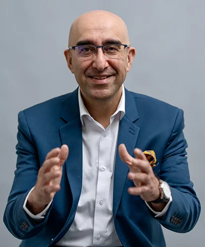 A friendly man with a bald head, glasses, and a blue suit smiles and gestures with his hands.