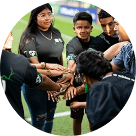 Several young male and female Hispanic children. They are standing in a circle on a soccer field and joining hands in the middle.