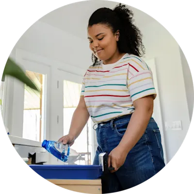 A young woman stands in a home kitchen. She is dropping a blue bottle into a trash drawer.