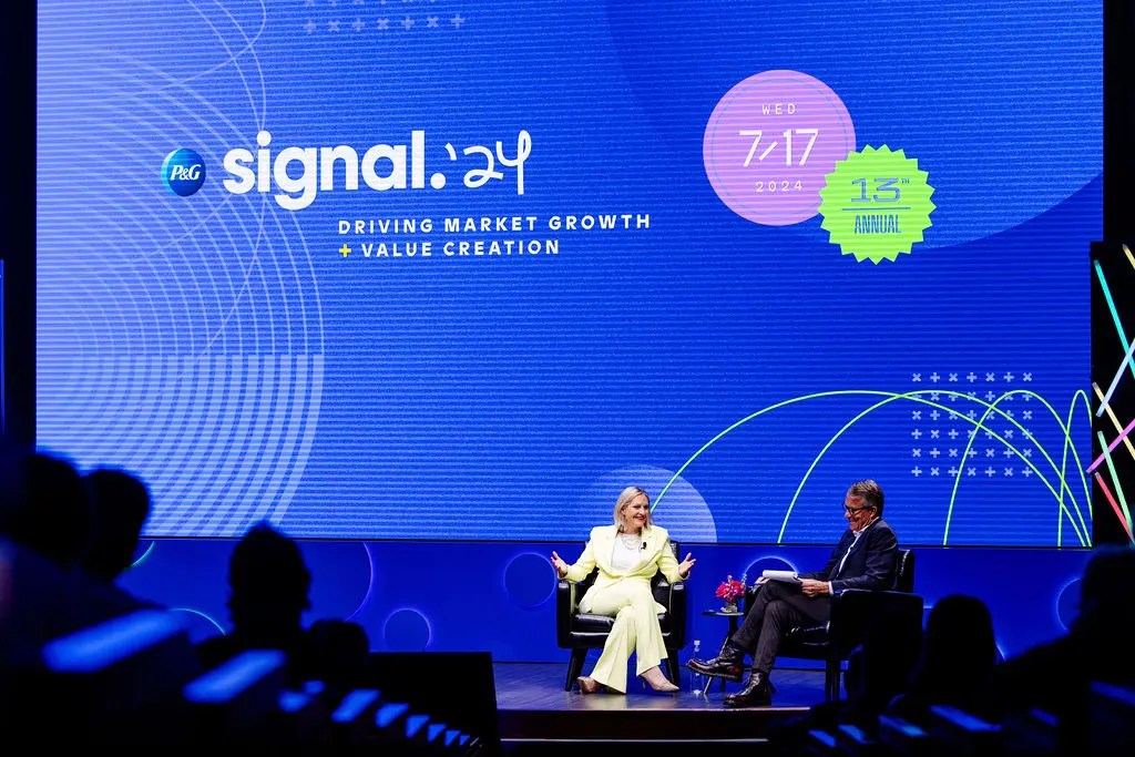 A woman with shoulder length blonde hair and a man in a blue suit sit and talk on a stage. A large blue graphic is displayed in the background with text that says "Signal '24. Driving market growth and value creation."