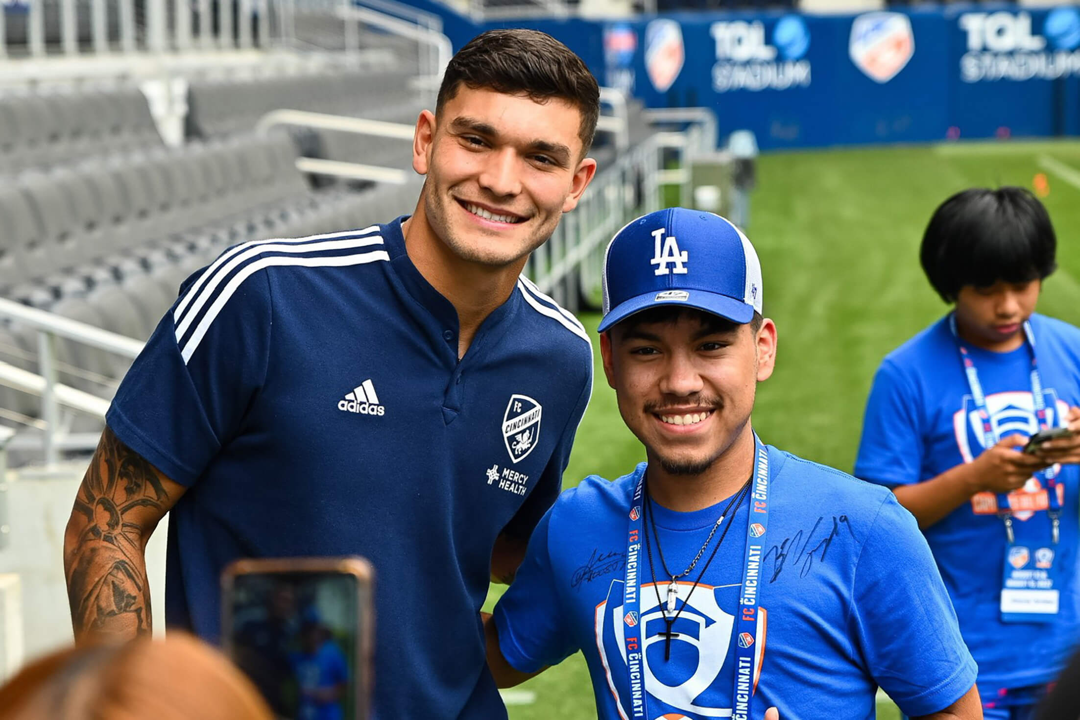 A Hispanic male in a dark blue soccer uniform. He stands next to a young Hispanic male student in a bright blue hat and t-shirt. They are standing on a soccer field.