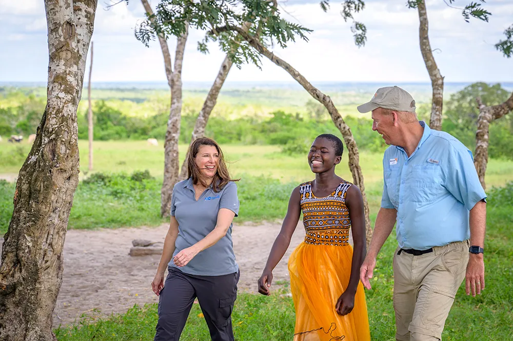 A woman and a man walk outdoors with a smiling teenage girl in a multicolor dress along a dirt path in a green rural landscape.