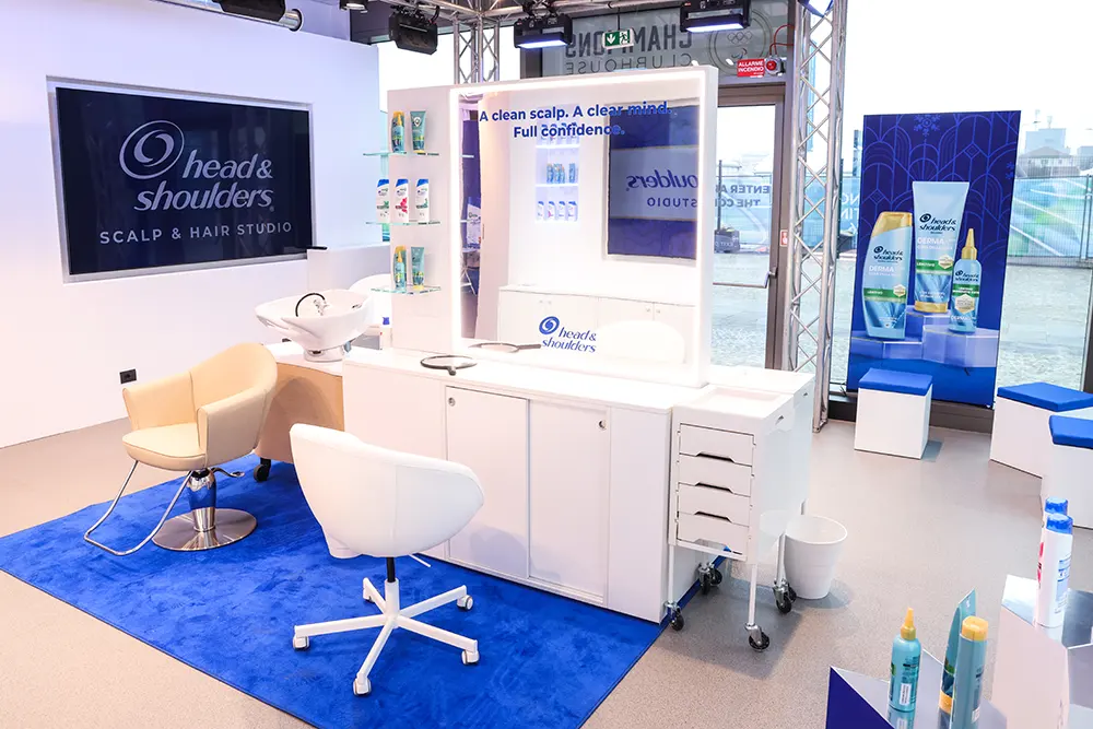 A Head & Shoulders Scalp & Hair Studio setup with a wash station and salon chair on a blue carpeted area. A large mirror surrounded by lights displays the wording "A clean scalp. A clean mind. Full Confidence." 