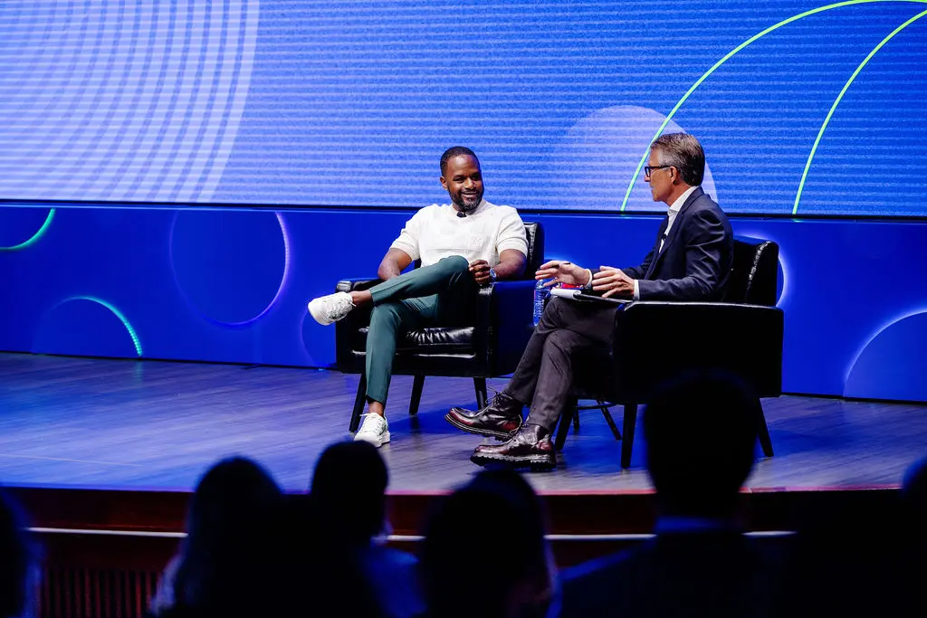 A black man in a white shirt and blue pants and a white man in a blue suit sit and talk on a stage. A large blue graphic is displayed in the background.