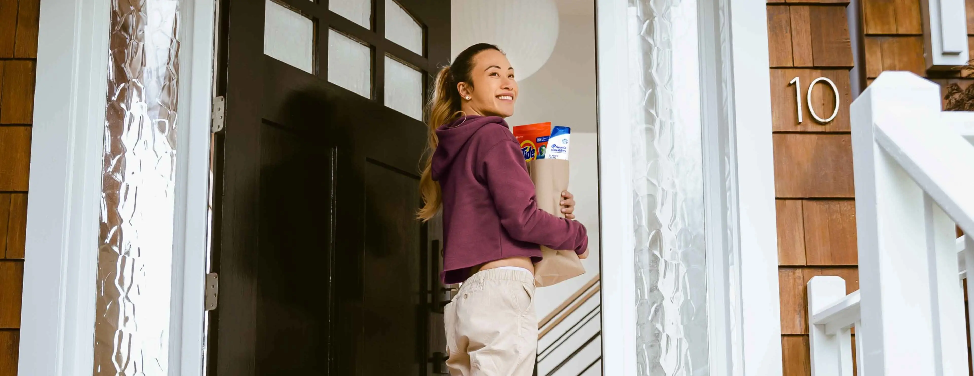 Woman enters front door of house while holding brown paper grocery bag containing Tide detergent and Head & Shoulders shampoo 