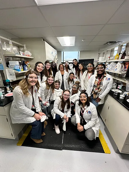 A group of young women in lab goggles and white lab coats post together in a group. They are in a laboratory.