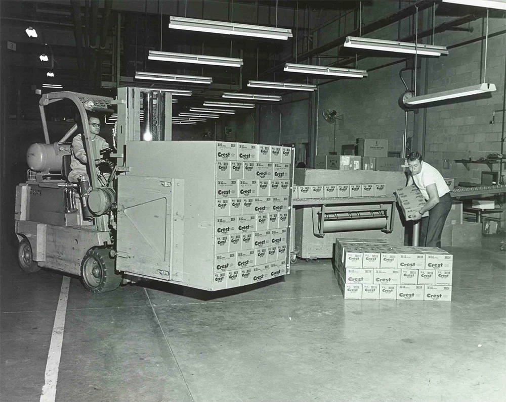 Inside a warehouse, a forklift operator transports pallets of Crest toothpaste boxes, while another worker handles individual boxes.