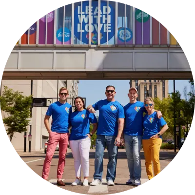 Five P&G employees in matching blue shirts smile and stand on the street under a bridge decorated with rainbow colors.