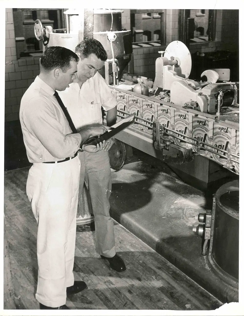 Black and white photo of two men examining a clipboard in front of a manufacturing line full of boxes of Tide.