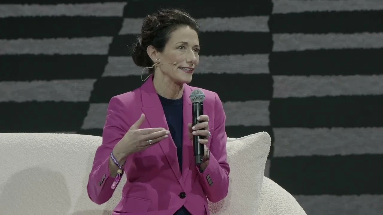 Woman in a bright pink pantsuit holding a microphone on stage and talking