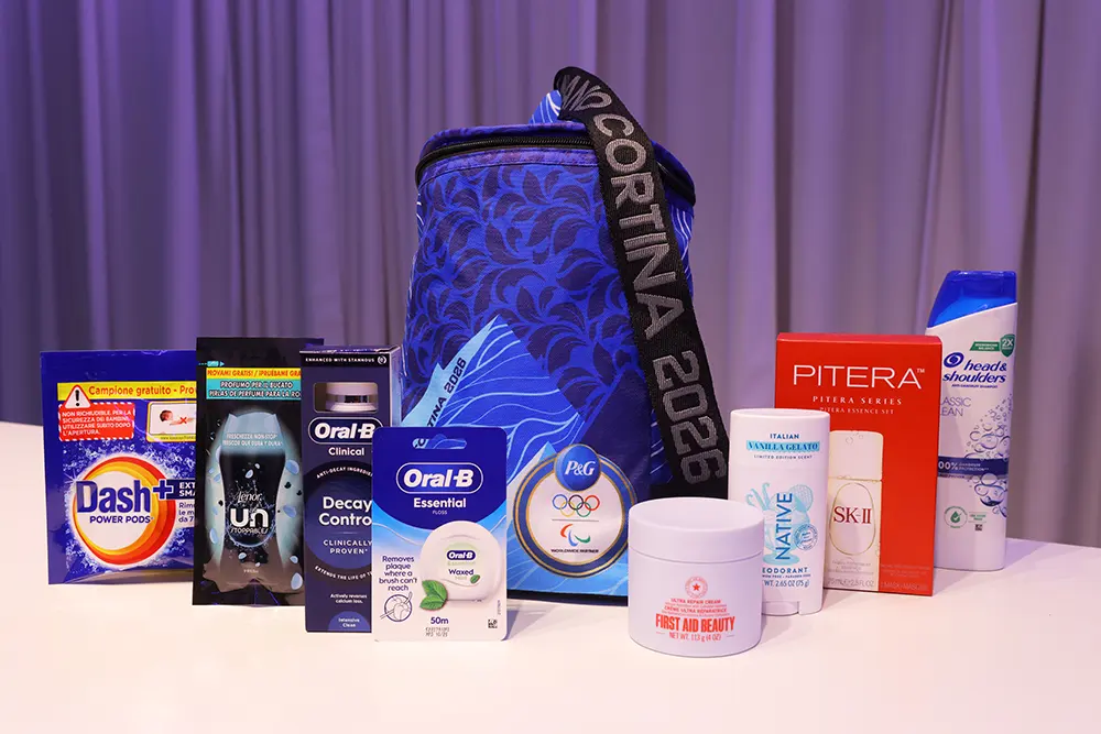 A collection of P&G products displayed on a white surface against a purple curtain backdrop. Products include a blue patterned bag with "Milano Cortina 2026" on the strap and the P&G Worldwide Olympic and Paralympic Partner medallion.