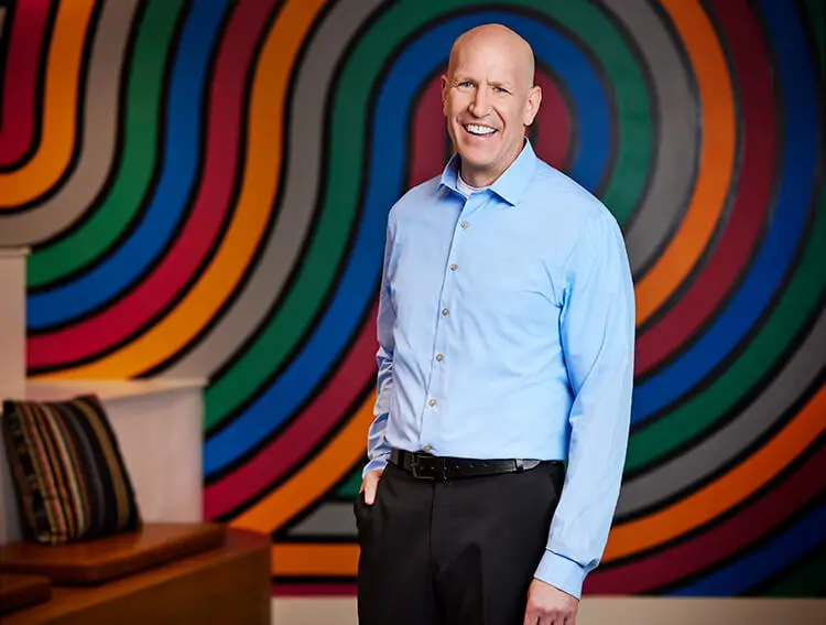 A tall, white male smiles. He is wearing black pants and a light blue, long sleeve shirt.