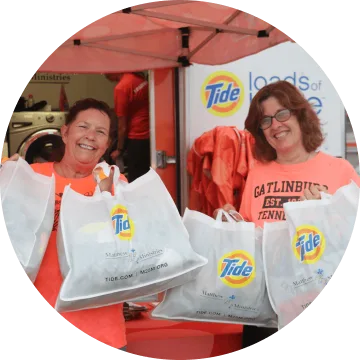 Two smiling women are holding two white tote bags with the Tide logo on it. They are both wearing orange t-shirts and standing under a tent. 