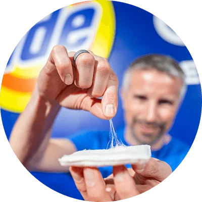 A man holds up a white square shaped swatch in one hand. With the other he is pinching and lifting white fibers from the swatch. The round yellow, orange and blue Tide logo is displayed on a bright blue backdrop.