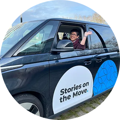 A woman with short dark hair and glasses waves from a dark van with a white and blue circular logo on the door that says, “Stories on the Move.”