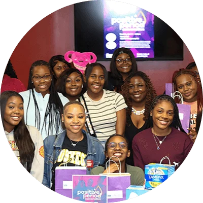 A group of seventeen young black college women gather and smile in front a table displaying Tampax period products and packaging.