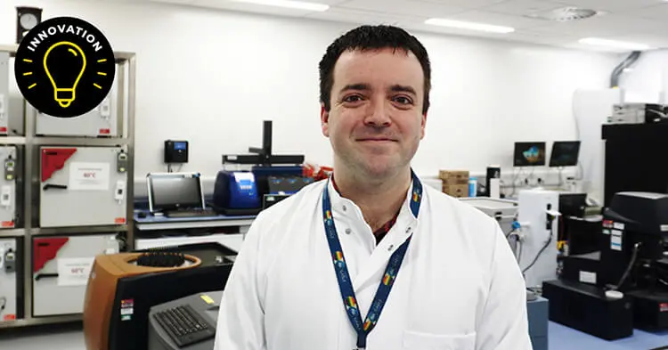 A man with short dark hair wears a white lab coat and a dark blue lanyard. He stands in front a digital lab, with keyboards, monitors and other devices in the background.