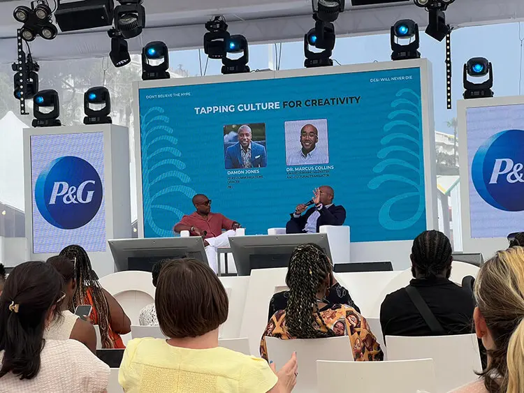 Two black men in business casual attire sit together on a stage as they engage in discussion. Their respective headshots are projected on a large screen behind them.