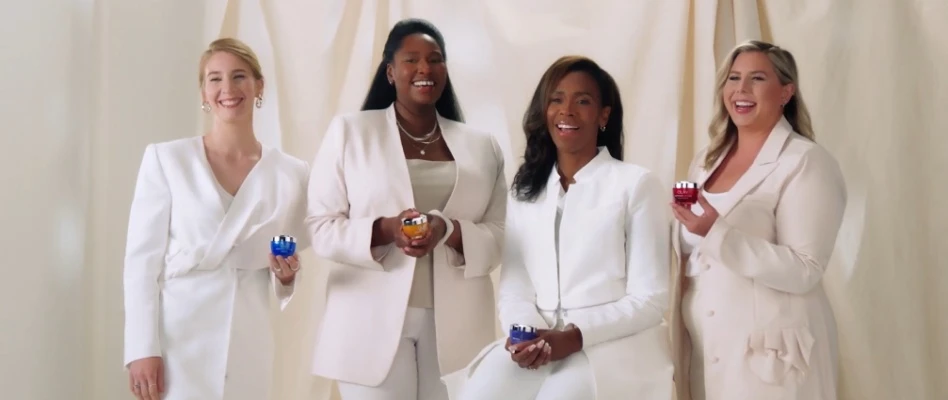 Four women in white suits smiling and looking towards the camera holding Olay cream products.