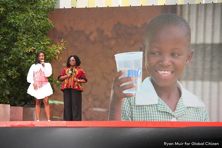 Two black women stand on a stage together with a screen featuring a black child holding a clean cup of water.