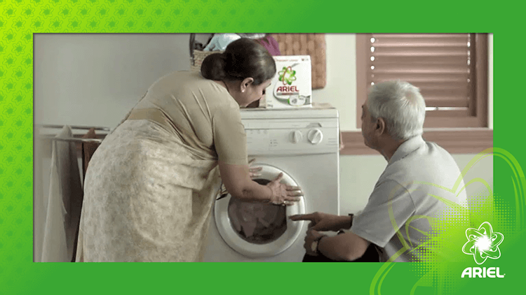 An Indian couple kneel in front of a white washing machine. A box of Ariel laundry detergent sits on top of the machine.