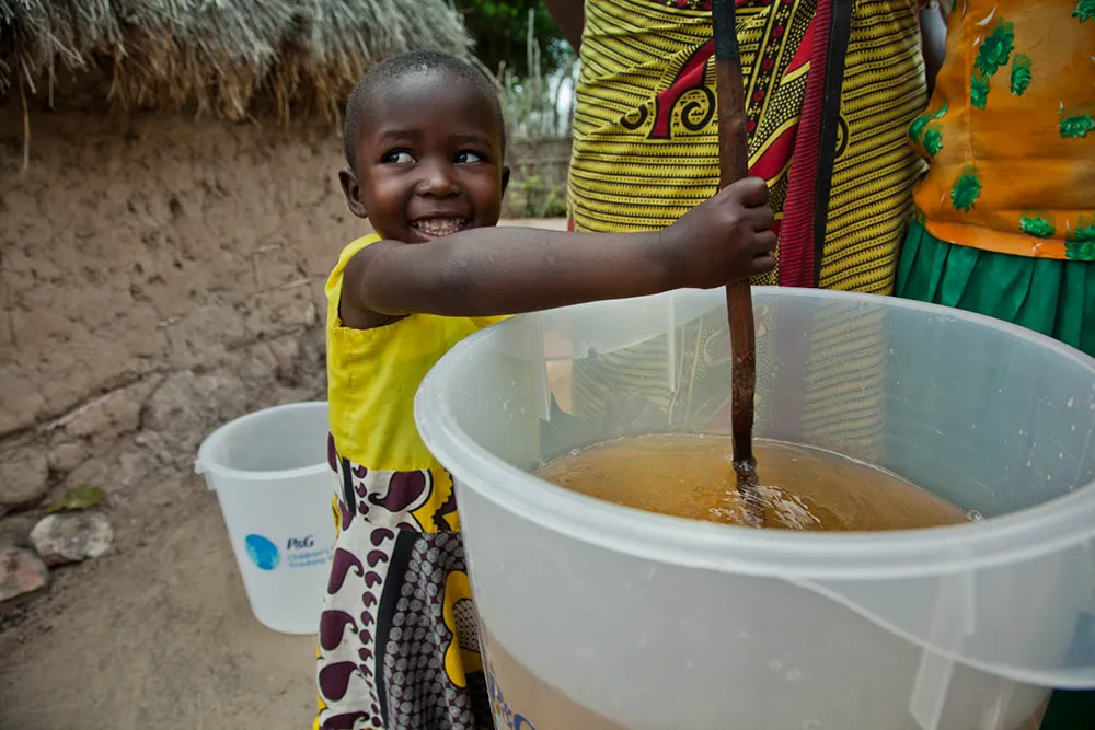 A young child stirs dirty water in a large clear bucket with a stick while an adult stands nearby.