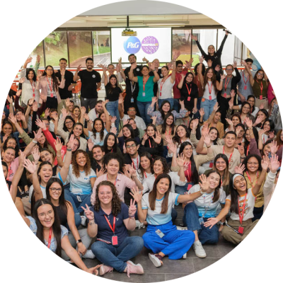 A large group of Hispanic Procter & Gamble employees pose, with many sitting on the floor and others standing in the background. They are waving and smiling. The group is predominantly female.