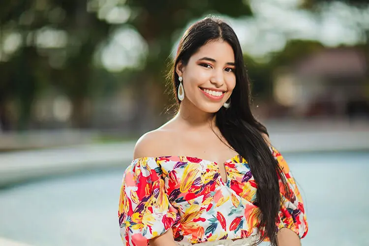 A young Hispanic woman with long dark hair, long white earrings and a brightly colored patterned blouse smiles and poses in an outdoor setting for a headshot.