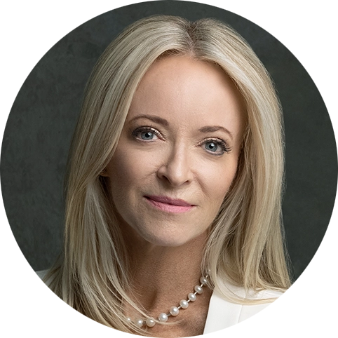 Professional headshot of Mindy Sherwood with shoulder-length blonde hair, wearing a white blazer and pearl necklace, facing the camera against a dark background.