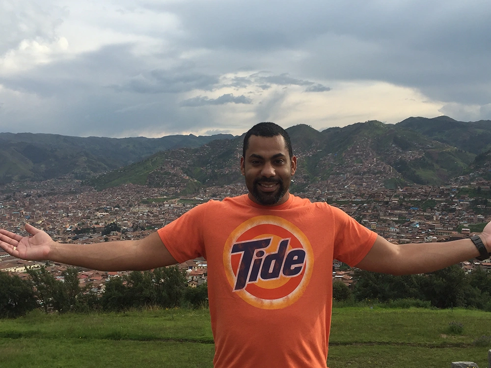 A smiling man in an orange Tide t-shirt opens his arms wide on a green hill overlooking a large valley, city and mountains.