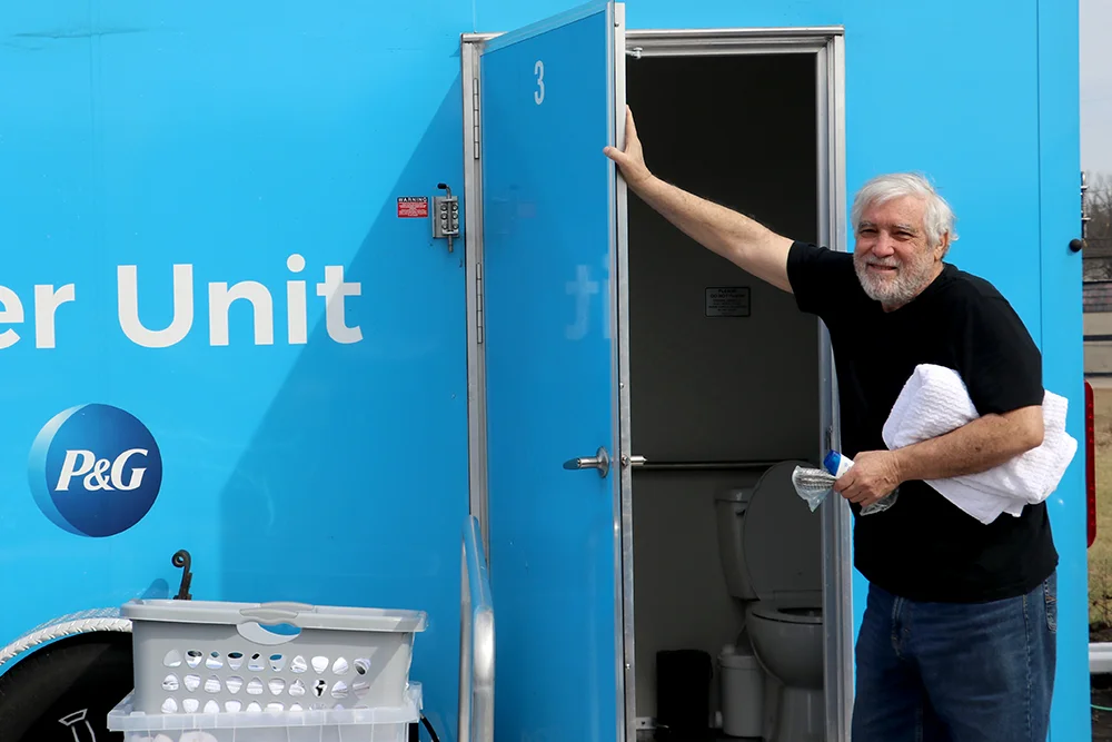 Person stands at the open door of a blue mobile unit labeled “Shower Unit” with a P&G logo, holding a folded white towel and a water bottle; a toilet is visible inside the unit.