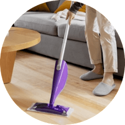 A person is cleaning the hardwood floors in a living room. They are only visible from the hip down, and they are using a purple Swiffer PowerMop.