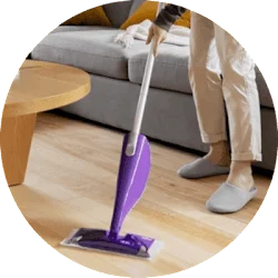 A person is cleaning the hardwood floors in a living room. They are only visible from the hip down, and they are using a purple Swiffer PowerMop.