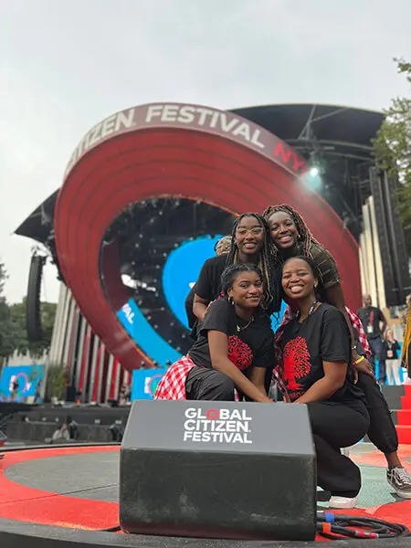 Four young Black women pose together. They're smiling and wearing matching black activewear outfits. A Global Citizenship sign is displayed in front of them.