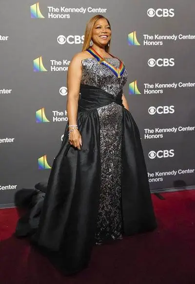 A black women wears a black and silver dress as she smiles on the red carpet. A black backdrop with multiples logos indicates the event is for The Kennedy Center Honors.