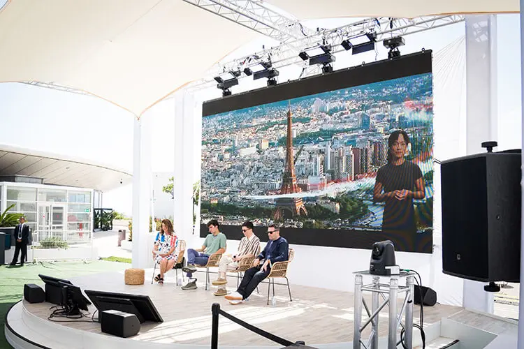 Three men and a woman sit and stand on a stage. An aerial image of Paris and Eiffel Tower is projected onto a large screen in the background.