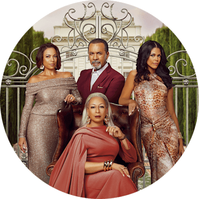 A woman in a long gown sits on a tall leather chair. Two women and a man in formal attire stand behind her. They all stand in front of an iron gate. Trees and a white mansion in the background.