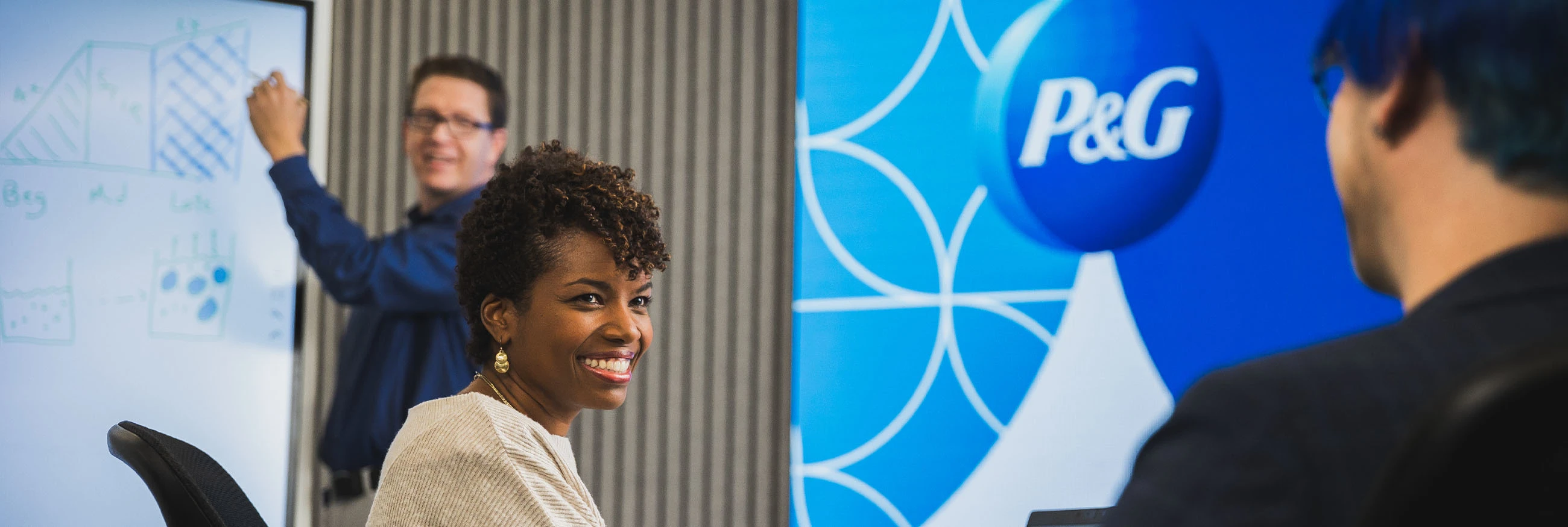 Three P&G employees work together – a man writing on whiteboard, a woman sitting at a table and smiling, and a third person with their back to the camera.  