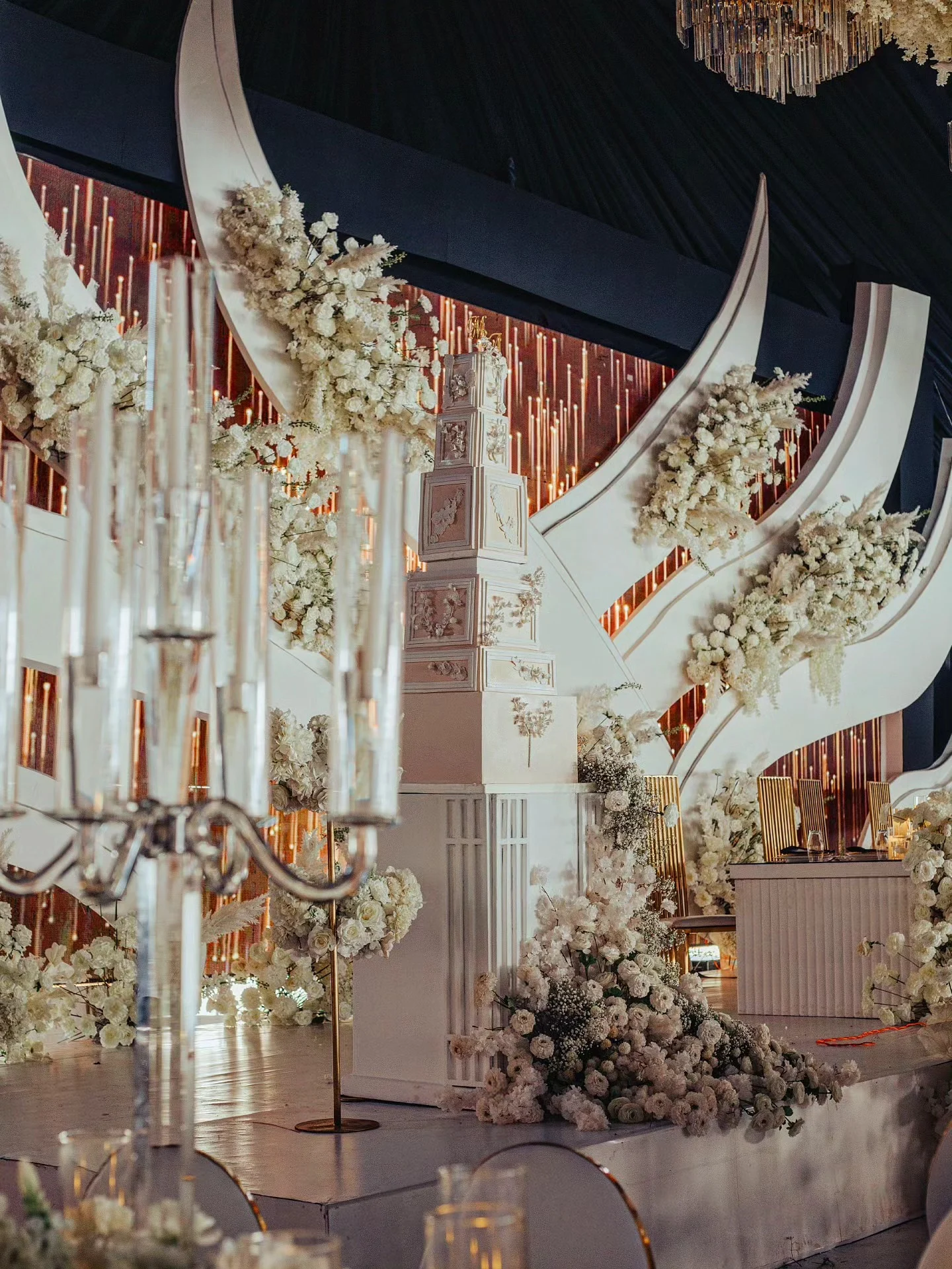 An ornate white wedding cake displayed amid white floral arrangements