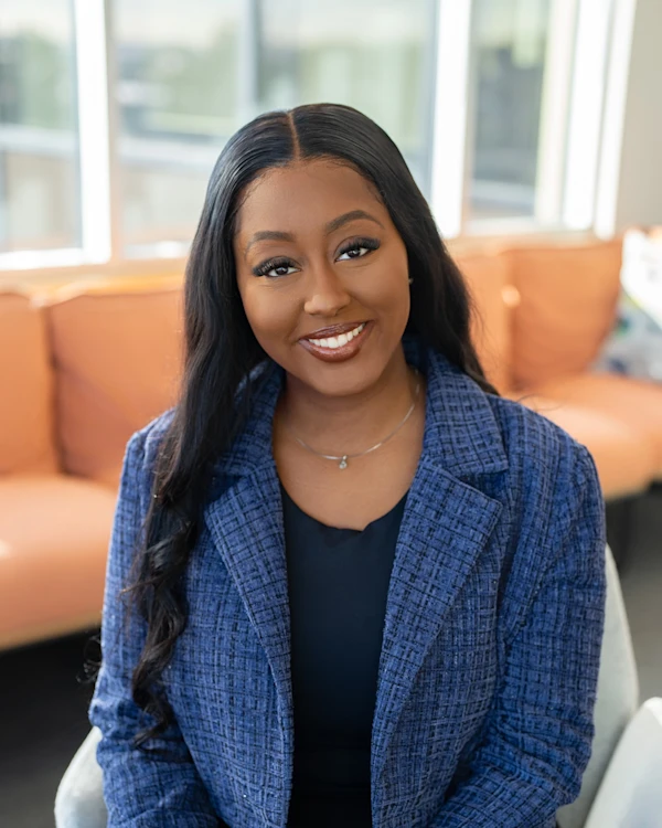 A woman wearing a blue blazer smiling with a window and orange sofa in the background.