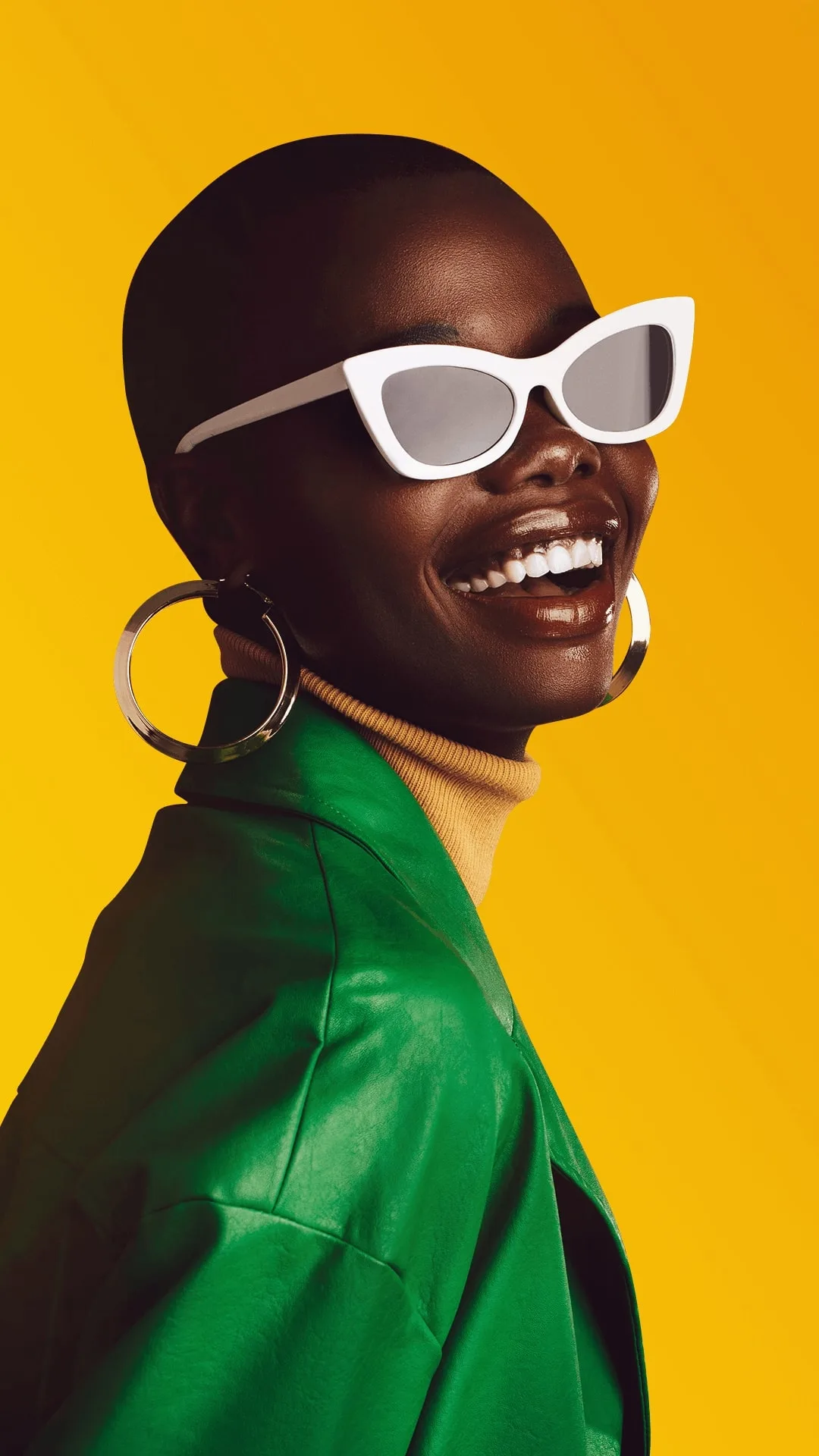 A smiling woman wearing large earrings and white sunglasses against a bright yellow background.