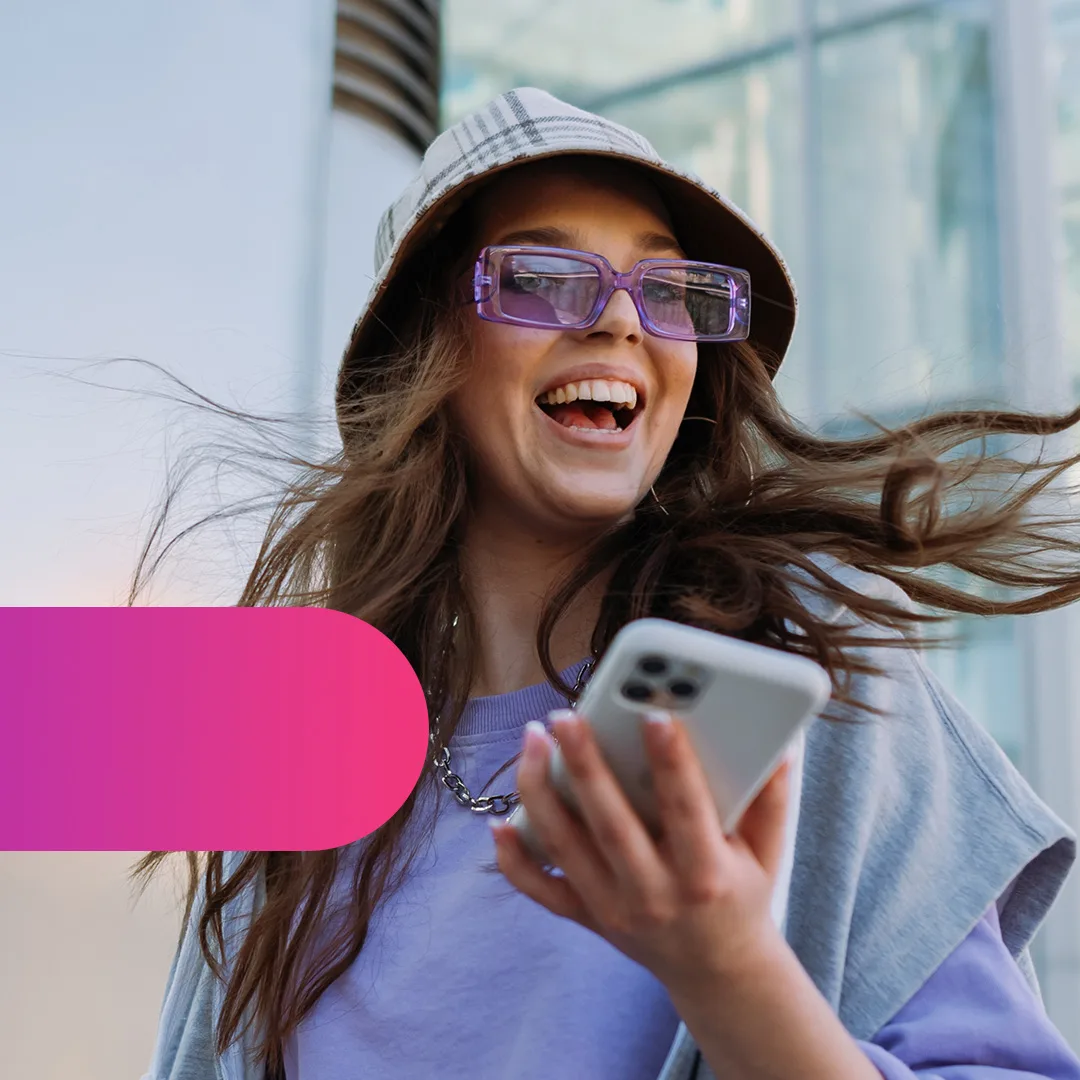 A smiling woman wearing sunglasses and a bucket hat holds a smartphone, with her hair flowing in the wind outdoors.