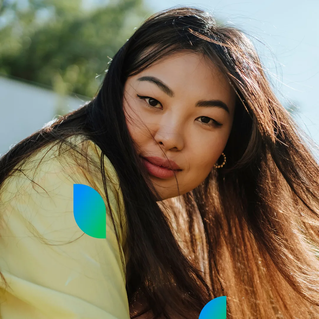 A woman with long, straight hair leans forward, her hair gently blowing in the breeze outdoors.
